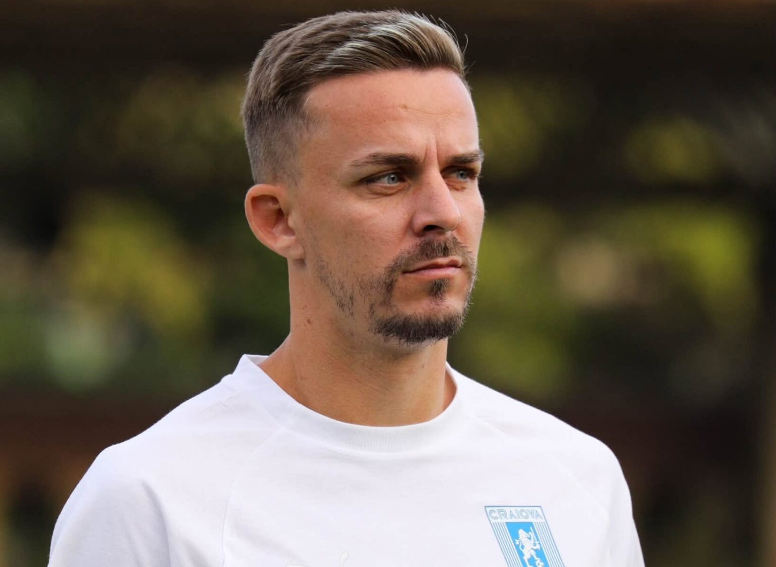 Portrait of a man in a white shirt with a blue crest on the chest, looking pensively to the right in an outdoor setting