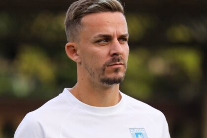 Portrait of a man in a white shirt with a blue crest on the chest, looking pensively to the right in an outdoor setting