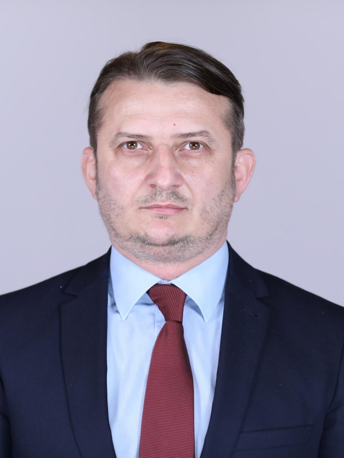 PecinginaGheorghe - Jurnalul Olteniei – Știri din Craiova și Oltenia Professional head-and-shoulders portrait of a man in a navy suit, light blue shirt, and red tie against a plain gray background.