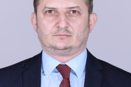 Professional head-and-shoulders portrait of a man in a navy suit, light blue shirt, and red tie against a plain gray background.