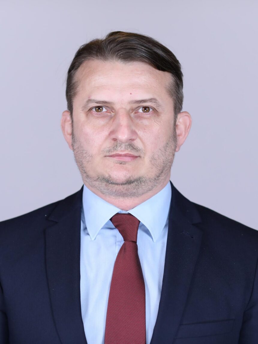 Professional head-and-shoulders portrait of a man in a navy suit, light blue shirt, and red tie against a plain gray background.