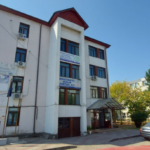 Three-story white building with banners and a glass entrance, cars parked in front under a blue sky isn’t