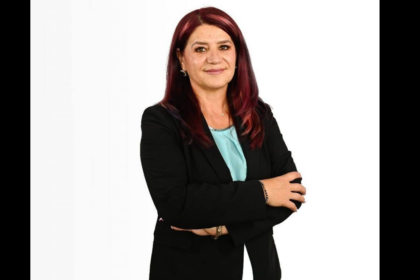 Professional woman with burgundy hair in a black blazer, arms crossed, wearing a light blue blouse, against a white background.