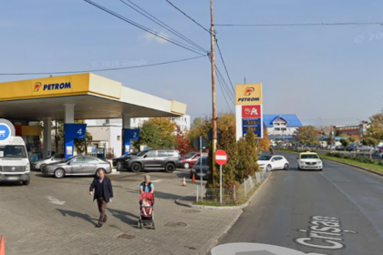 Petrom gas station with blue pump islands, several cars at the pumps, and pedestrians including a woman pushing a stroller on the sidewalk.