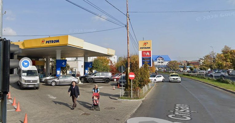 Petrom gas station with blue pump islands, several cars at the pumps, and pedestrians including a woman pushing a stroller on the sidewalk.