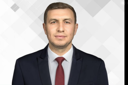 Professional headshot: man in dark suit and red tie against a light geometric background, facing camera