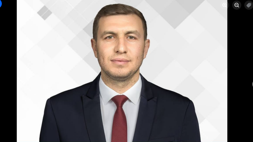 Professional headshot: man in dark suit and red tie against a light geometric background, facing camera