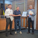 Three men in a formal room, two holding certificates during an award presentation; a Ukrainian flag and a framed photo are visible in the background.