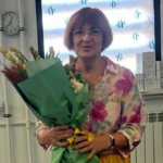 Woman in a floral blouse holding a large bouquet wrapped in green paper indoors.
