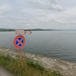 No parking sign by a concrete seawall along a calm lake or sea, with cloudy sky in the distance and a grassy roadside.