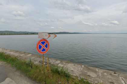 No parking sign by a concrete seawall along a calm lake or sea, with cloudy sky in the distance and a grassy roadside.
