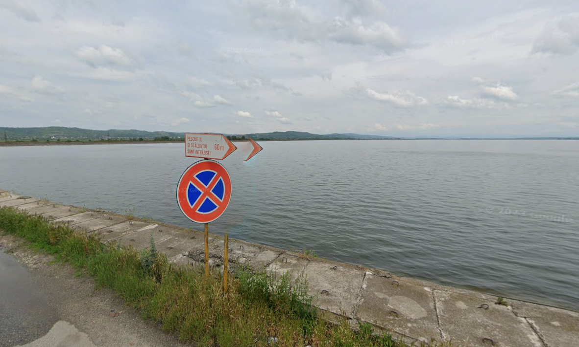 Screenshot 2026-09-23 093912 - Jurnalul Olteniei – Știri din Craiova și Oltenia No parking sign by a concrete seawall along a calm lake or sea, with cloudy sky in the distance and a grassy roadside.