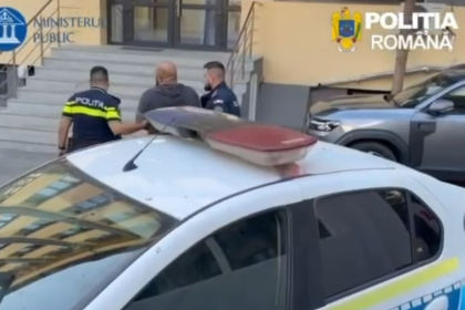 Two Romanian police officers accompany a man near a police car outside a government building with stairs in the background.