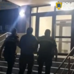 Three men walking up an outdoor staircase toward a glass door at night, railing on both sides.