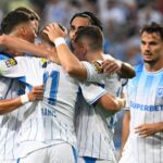 Soccer players in white-and-blue striped jerseys celebrate with a group hug on the field; one teammate watches nearby.