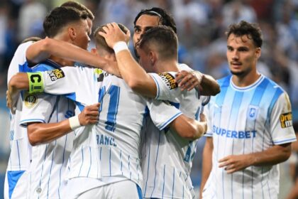 Soccer players in white-and-blue striped jerseys celebrate with a group hug on the field; one teammate watches nearby.