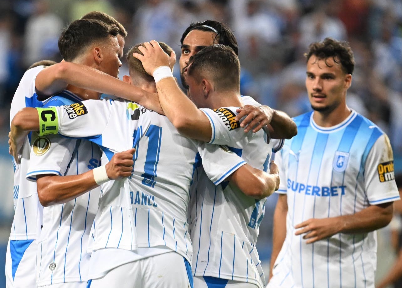Soccer players in white-and-blue striped jerseys celebrate with a group hug on the field; one teammate watches nearby.