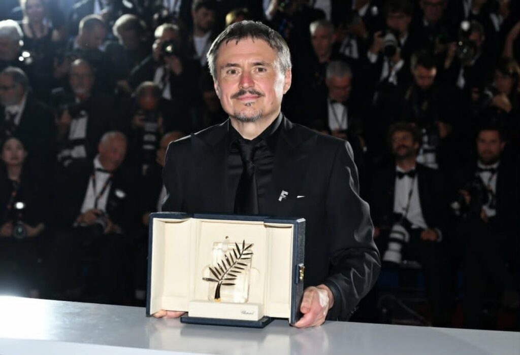 Man in a black suit and bow tie presents a framed award with a palm leaf, at a formal ceremony with photographers in the background/flashbulbs.