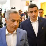 Two men in suits walk through a crowded room with photographers taking pictures behind them at a press event.