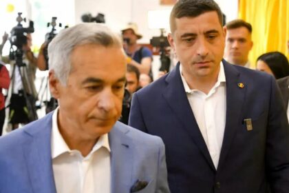 Two men in suits walk through a crowded room with photographers taking pictures behind them at a press event.