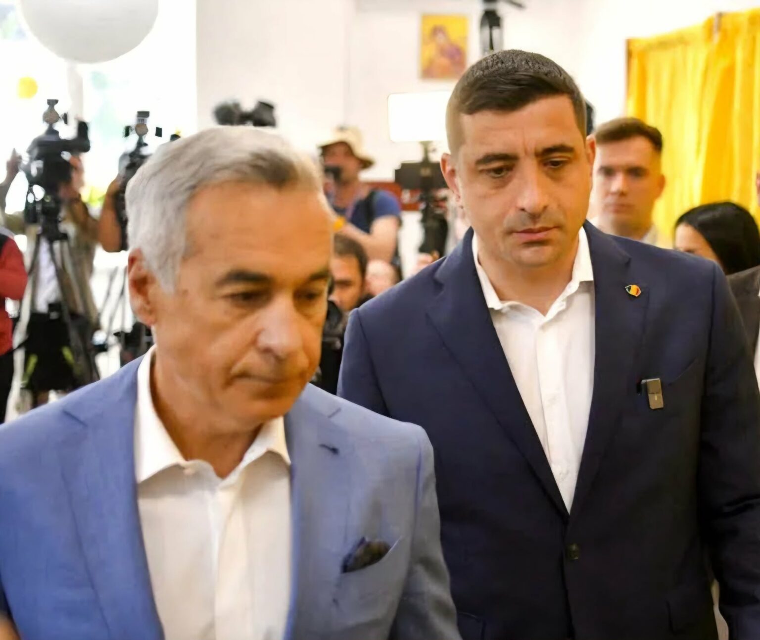Two men in suits walk through a crowded room with photographers taking pictures behind them at a press event.