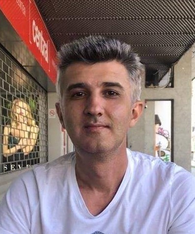 WhatsApp-Image-2023-09-19-at-114146 - Jurnalul Olteniei – Știri din Craiova și Oltenia Man with short gray hair wearing a white T-shirt sits at an outdoor cafe on a city street, with a storefront window and red sign behind him.