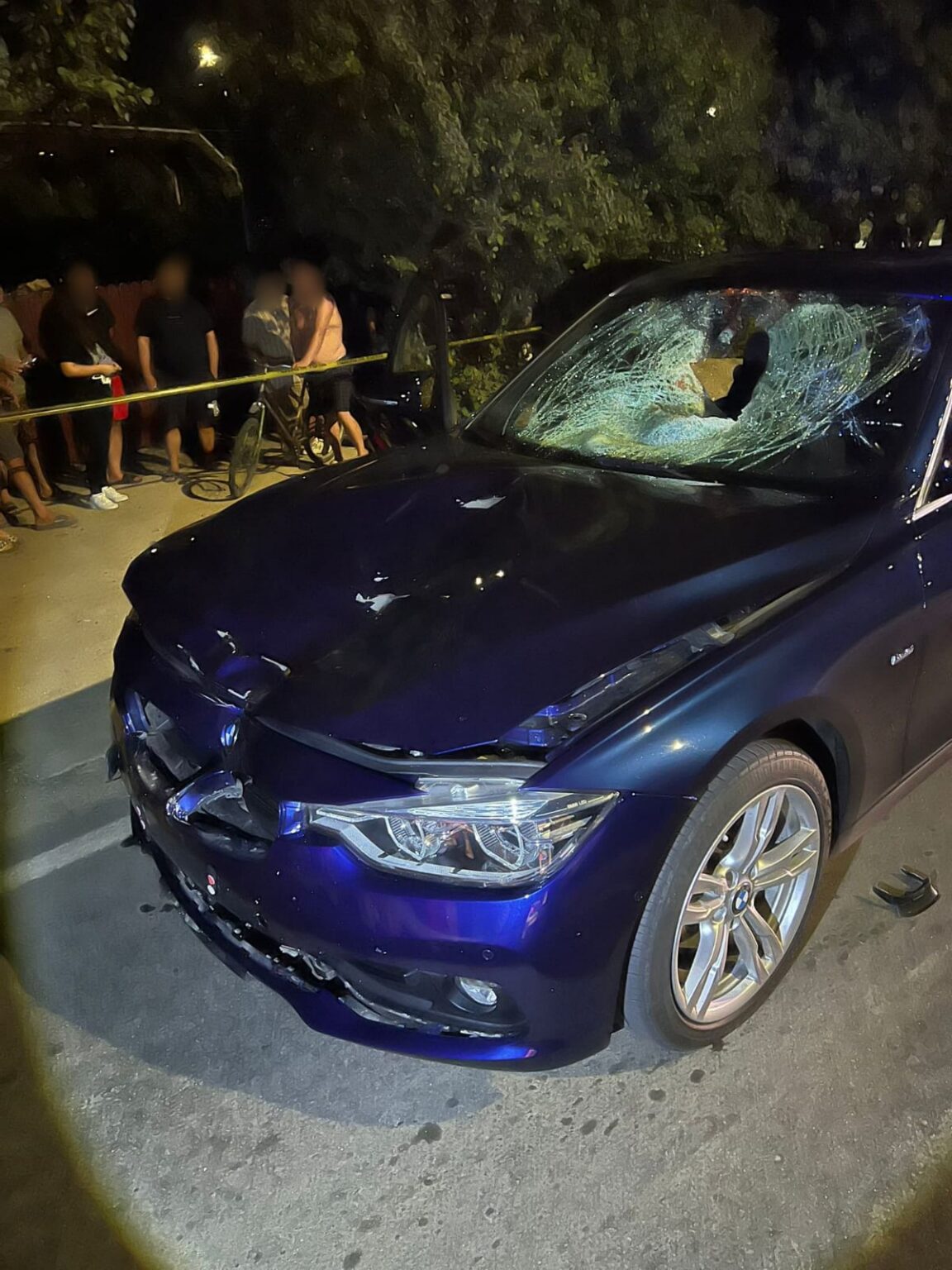 WhatsApp Image 2026-09-01 at 100742 - Jurnalul Olteniei – Știri din Craiova și Oltenia Blue sedan with extensive front-end damage and a shattered windshield at night, onlookers behind yellow caution tape nearby