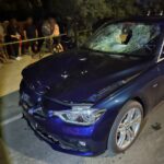 Blue sedan with extensive front-end damage and a shattered windshield at night, onlookers behind yellow caution tape nearby