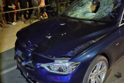 Blue sedan with extensive front-end damage and a shattered windshield at night, onlookers behind yellow caution tape nearby