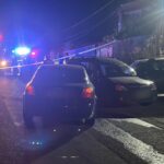 Nighttime street scene with flashing red and blue emergency lights, yellow tape cordoning off the area, and parked cars along the road outside a house.