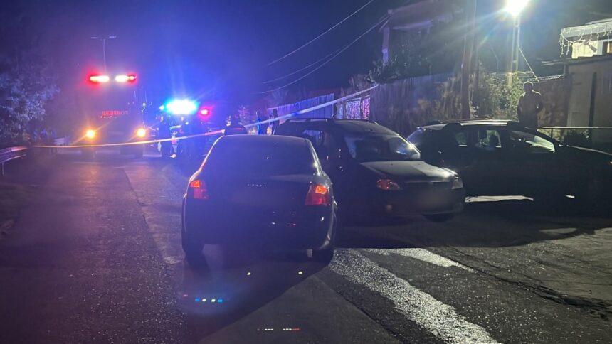 Nighttime street scene with flashing red and blue emergency lights, yellow tape cordoning off the area, and parked cars along the road outside a house.