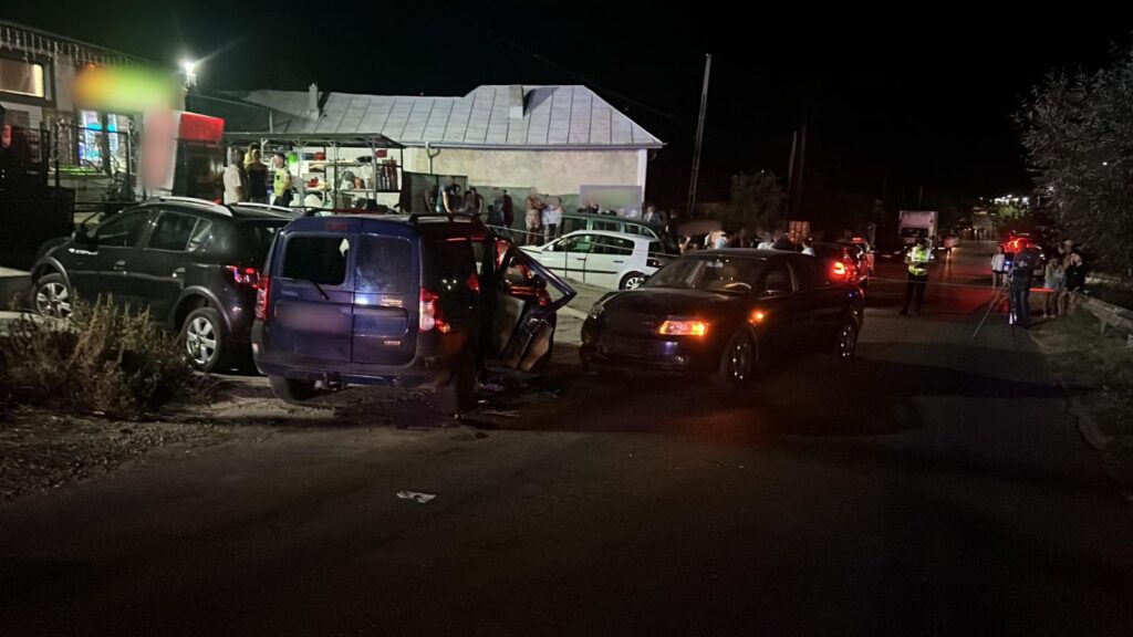 Nighttime street crash scene with a blue minivan door open, a dark sedan nearby, and bystanders and responders near a storefront building.
