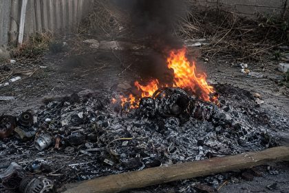 A pile of burning trash and debris in an outdoor alley, with orange flames, black smoke, and scorched rubble scattered on the ground.
