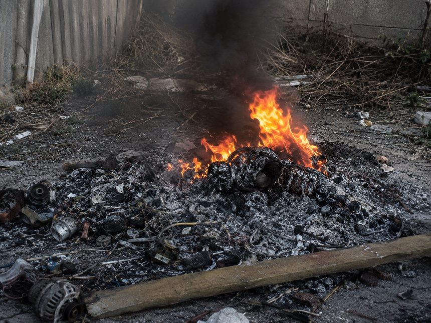 A pile of burning trash and debris in an outdoor alley, with orange flames, black smoke, and scorched rubble scattered on the ground.