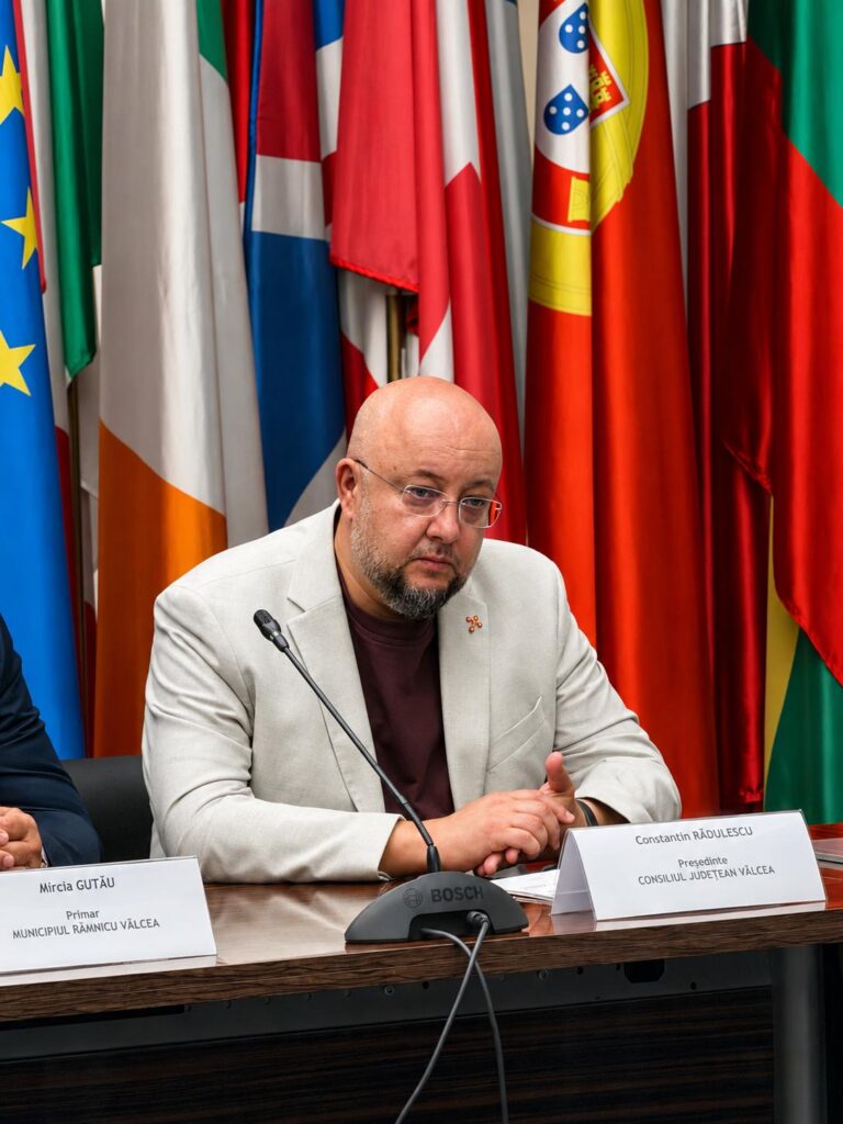 Bald man with glasses and a beard in a light gray suit speaks at a panel, with multiple national flags in the background.