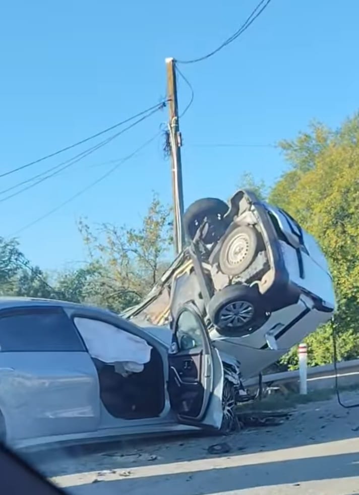 Two cars involved in a highway crash; a white SUV is perched on top of a gray sedan with both doors open and debris around them.