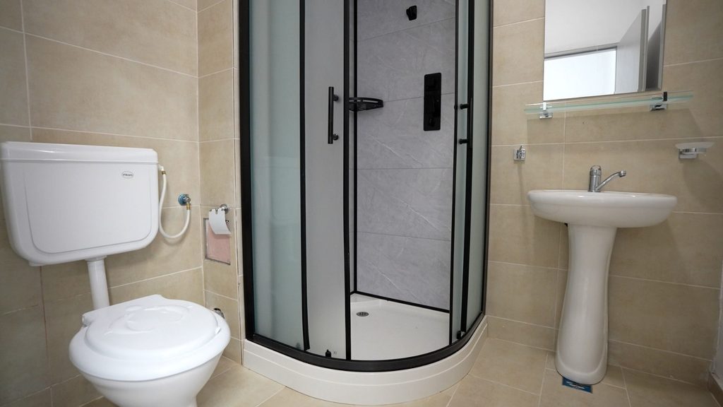 WhatsApp Image 2026-09-07 at 123847 - Jurnalul Olteniei – Știri din Craiova și Oltenia Bathroom with a curved corner shower enclosure, white toilet, and white pedestal sink on beige tiles.