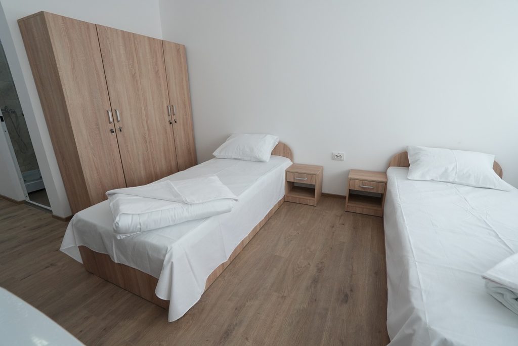 WhatsApp Image 2026-09-07 at 123849 - Jurnalul Olteniei – Știri din Craiova și Oltenia Two single beds with white linens and pillows in a bright minimalist bedroom, separated by small wooden nightstands and a large wooden wardrobe against the wall.