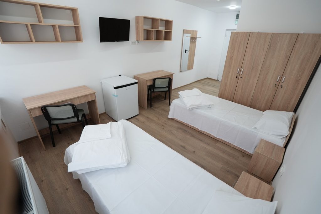 WhatsApp Image 2026-09-07 at 123850 - Jurnalul Olteniei – Știri din Craiova și Oltenia A clean dorm room with two single beds, white linens, wooden desks and matching wardrobes on a light wood floor.