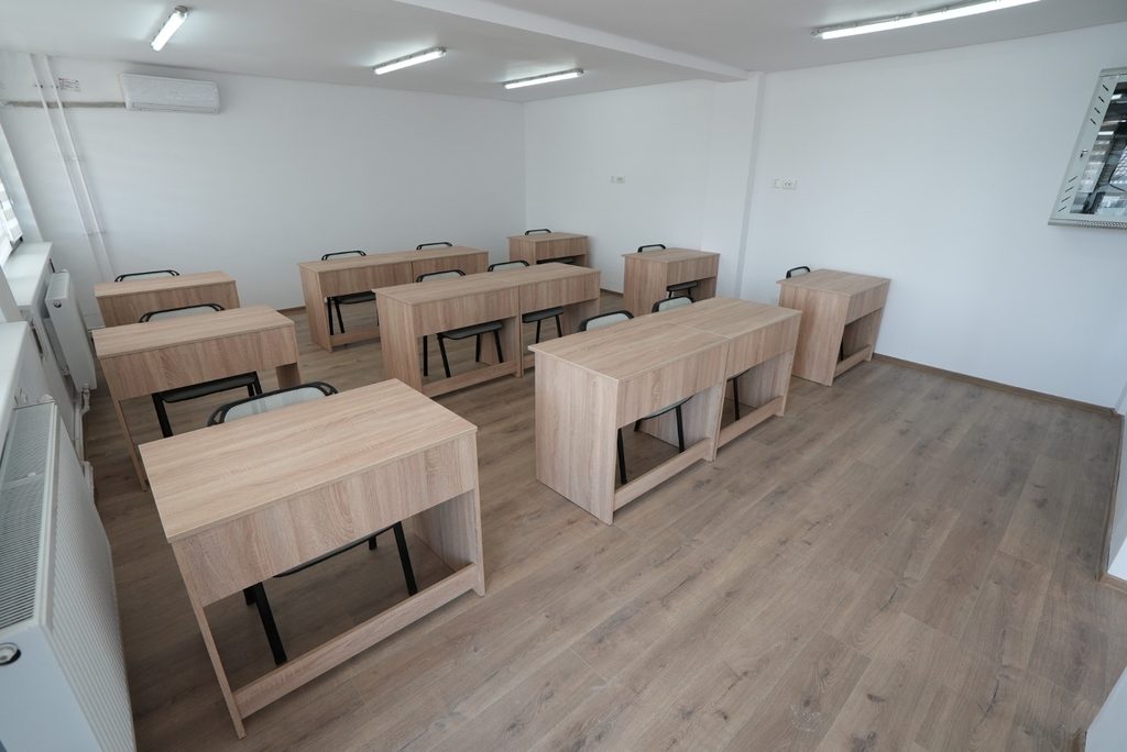 WhatsApp Image 2026-09-07 at 123851 - Jurnalul Olteniei – Știri din Craiova și Oltenia Empty classroom with light wood desks and black chairs arranged in rows on a light wood floor, white walls, and bright overhead fluorescent lighting.