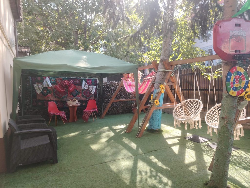 Backyard with a green canopy shelter over a seating area, red chairs, and colorful textiles under the shade.