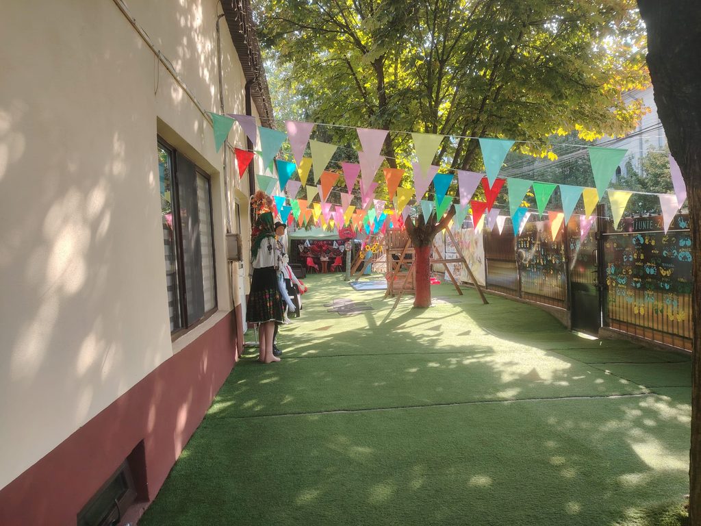 Outdoor courtyard with colorful triangular bunting overhead, sunlight filtering through trees and mannequins in traditional dresses nearby.