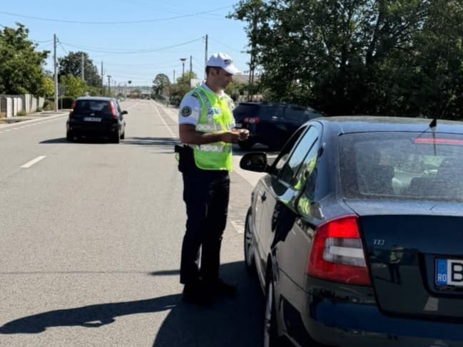 Traffic police officer in a high-visibility vest checks a driver beside a dark blue sedan on a sunny street, holding a notebook or device.