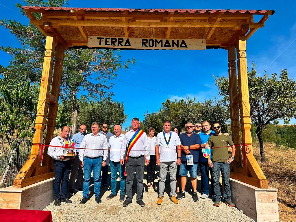 Group of men posing under a wooden arch reading 'TERRA ROMANA' at an outdoor ceremony, with red ribbon and a clear blue sky in the background.
