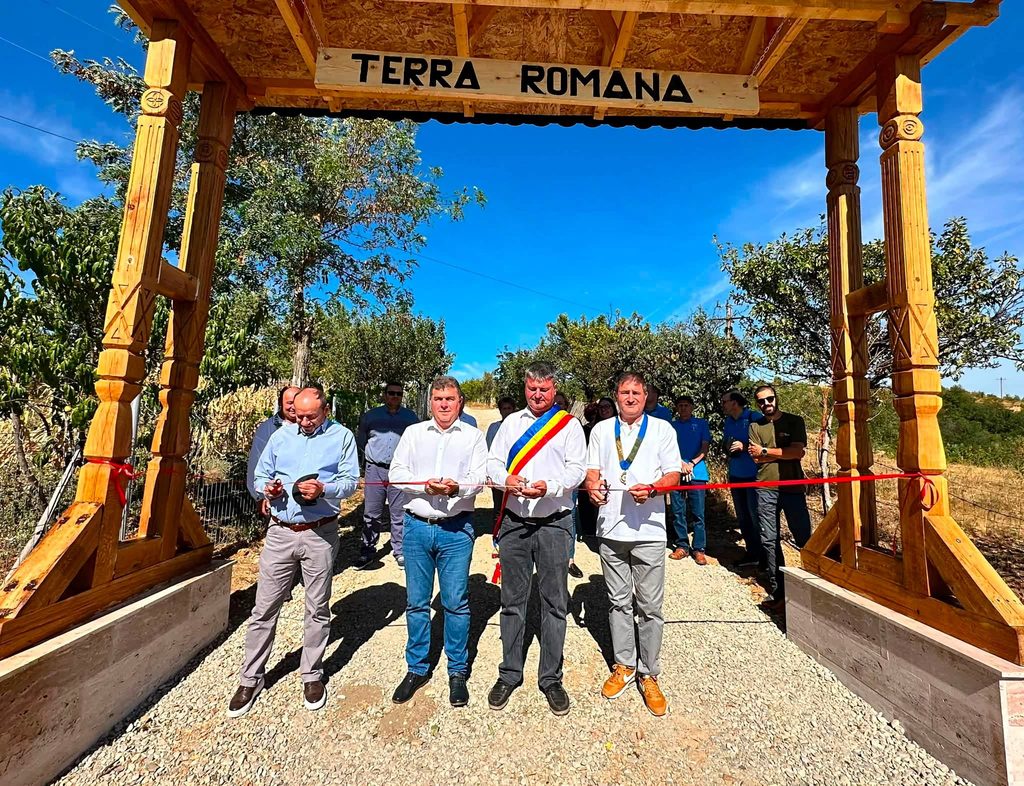 Group of people in a ribbon-cutting ceremony under a wooden arch with 'Terra Romana' sign on a sunny day outdoors.