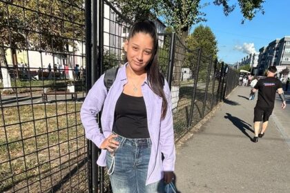 Young woman standing on a sunny sidewalk beside a black metal fence, wearing a purple shirt over a black top and blue jeans, carrying a backpack and a blue shopping bag.