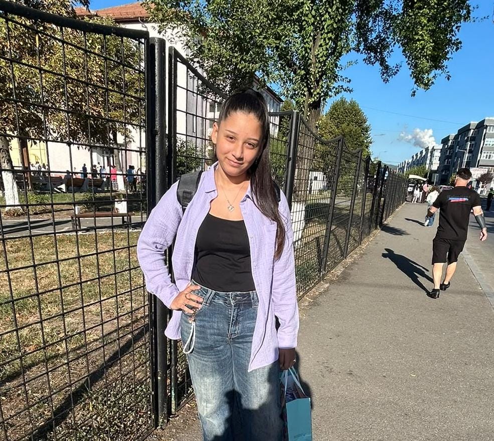 Young woman standing on a sunny sidewalk beside a black metal fence, wearing a purple shirt over a black top and blue jeans, carrying a backpack and a blue shopping bag.