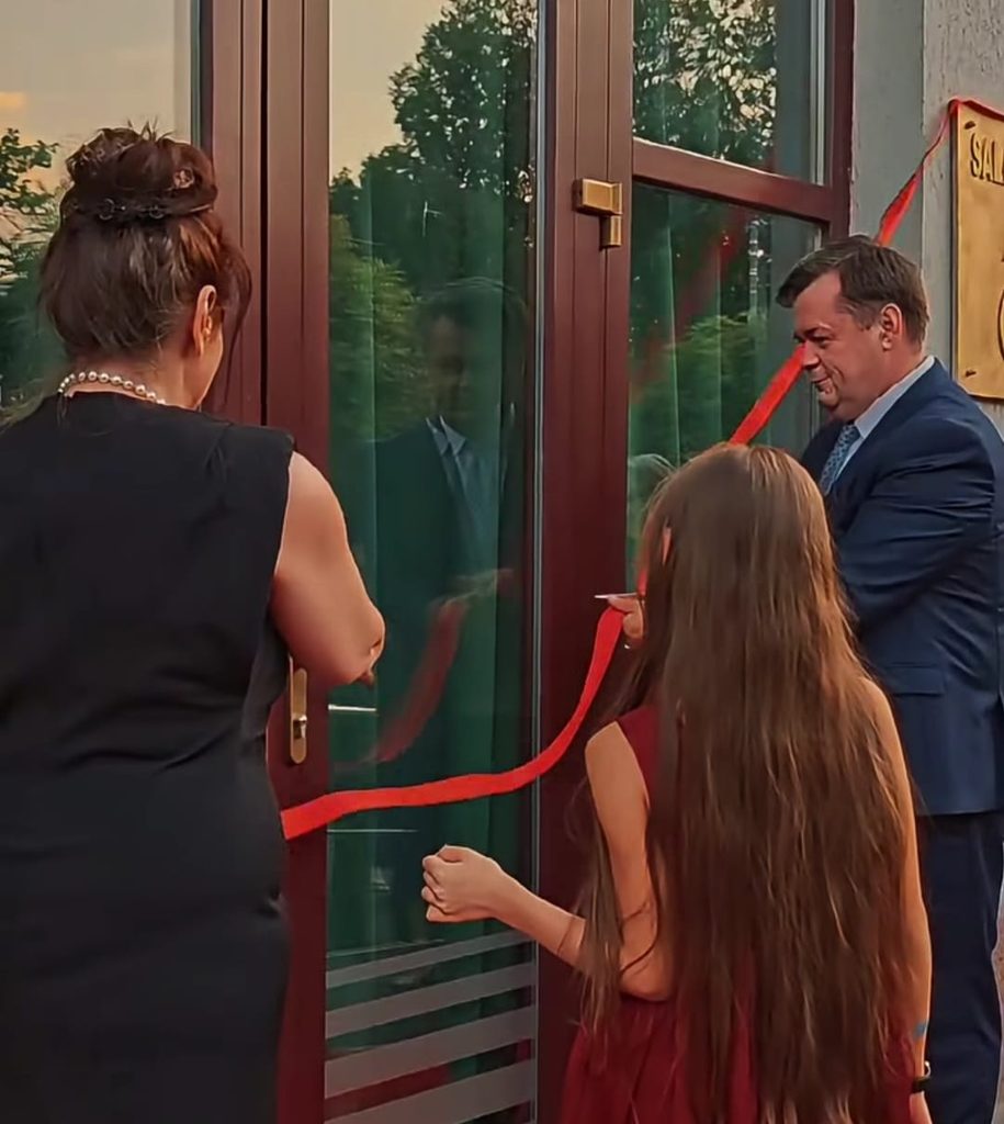 WhatsApp Image 2026-09-21 at 021016 1 - Jurnalul Olteniei – Știri din Craiova și Oltenia People cutting a red ribbon across glass doors at a storefront opening ceremony, with a man in a suit smiling on the right and a woman in black on the left.