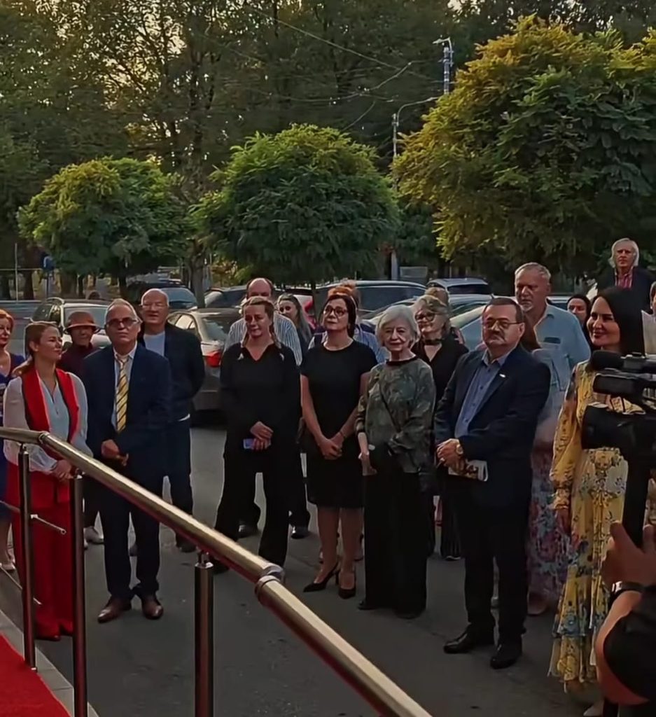 WhatsApp Image 2026-09-21 at 021016 2 - Jurnalul Olteniei – Știri din Craiova și Oltenia Group of formally dressed people standing on a sidewalk near a red carpet outdoors, with trees and parked cars in the background.