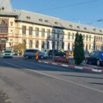 Historic city street with an ornate European-style building, multiple cars, and orange construction barriers under a clear blue sky.